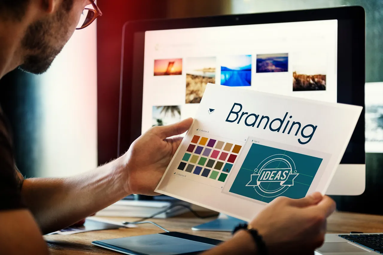 Person holding a printed branding concept beside a laptop and design materials