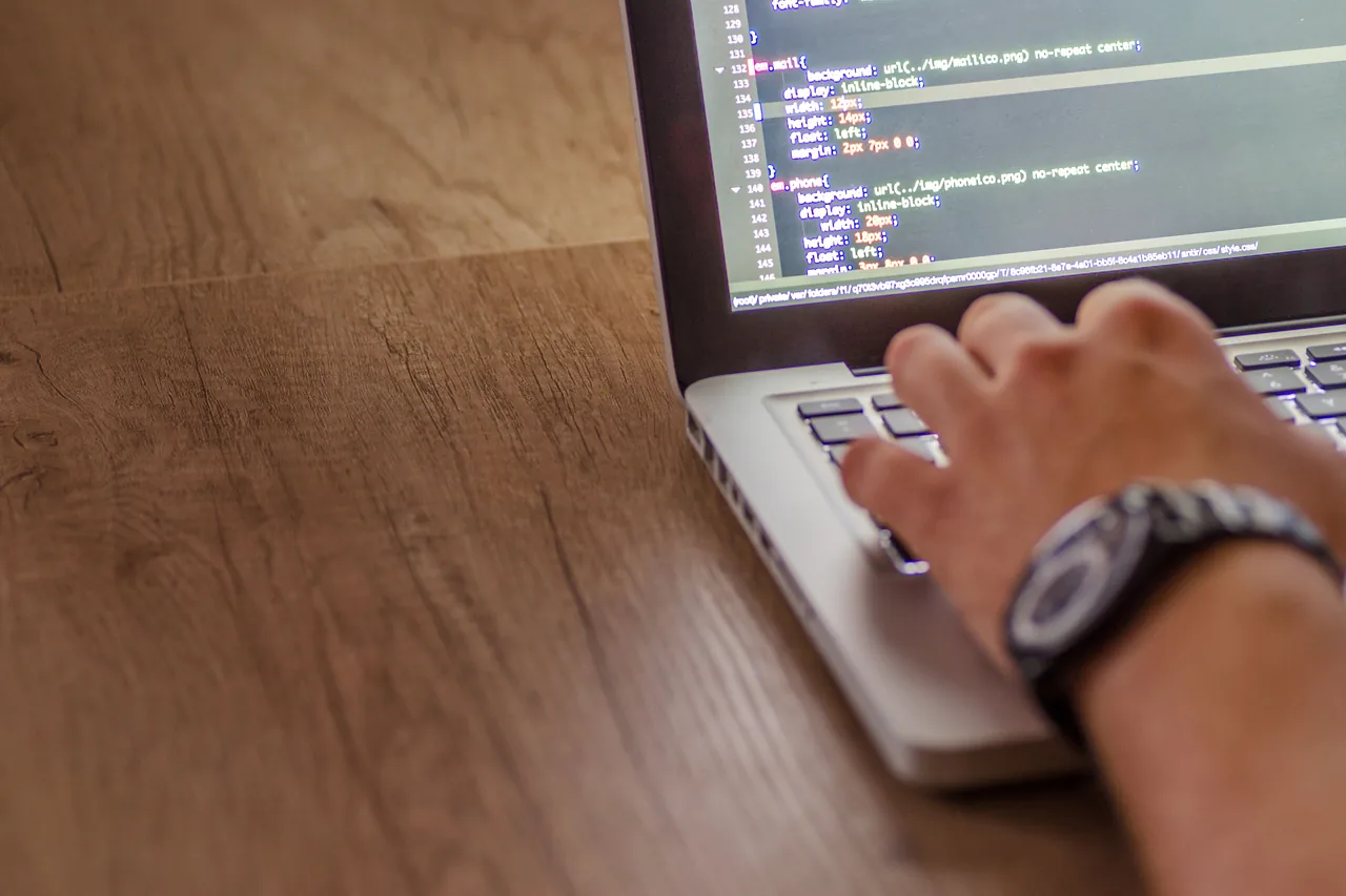 Hands typing on a laptop displaying HTML and CSS code on a wooden desk