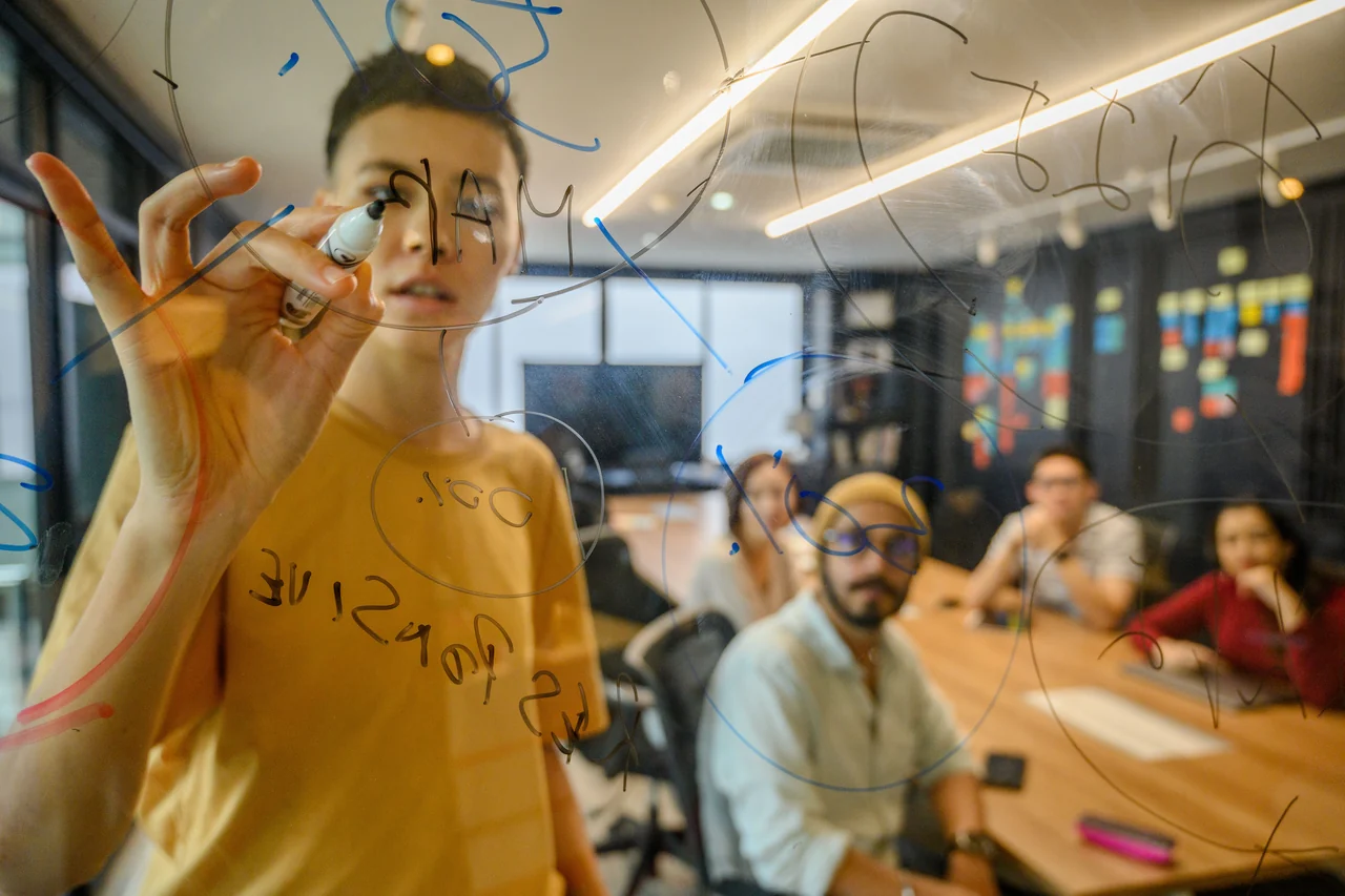 Person writing on a glass wall with four people seated around a conference table behind them