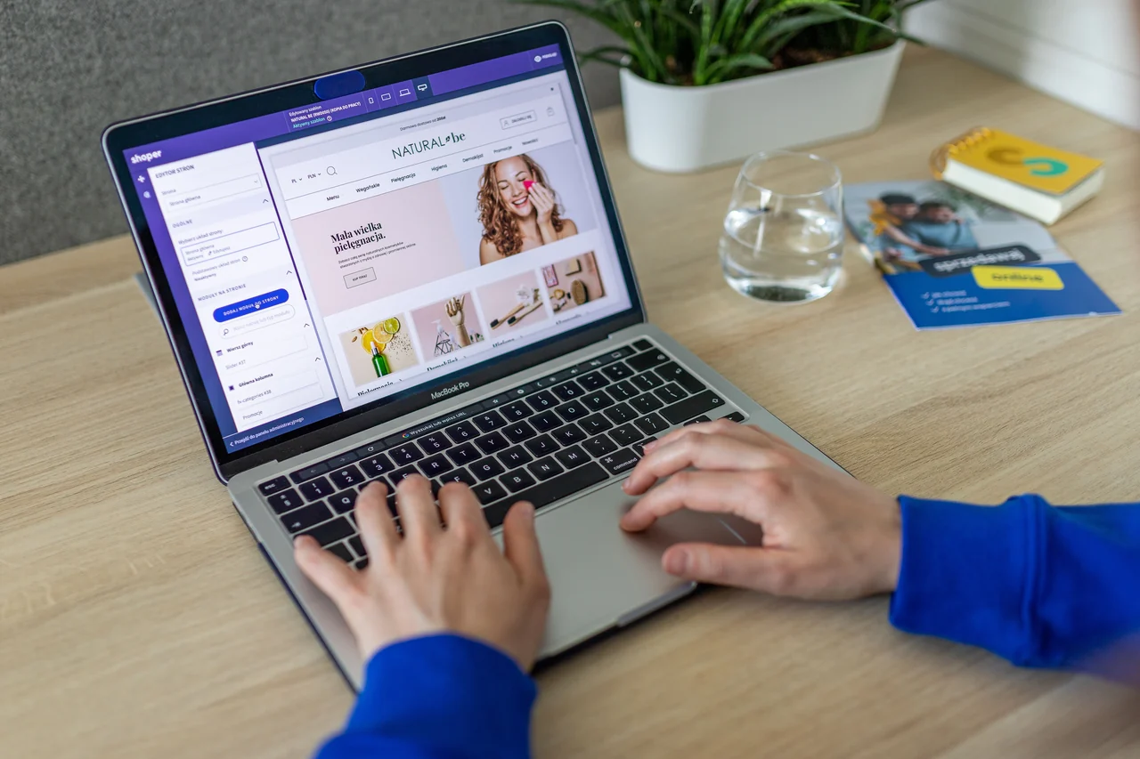 Hands using a laptop that displays a website editor beside a glass of water and a potted plant