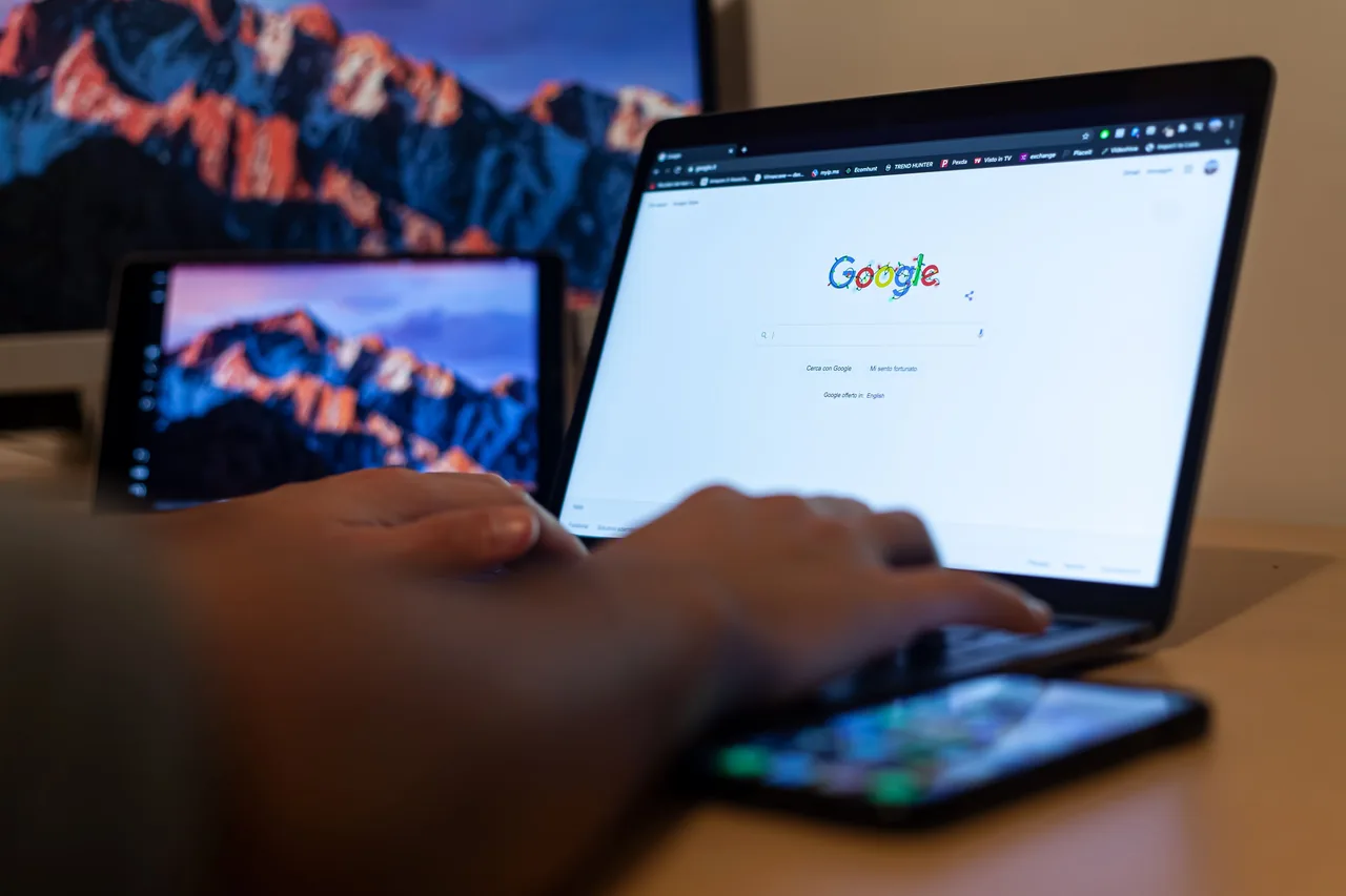 Hands typing on a laptop displaying Google Search, with a tablet and smartphone nearby